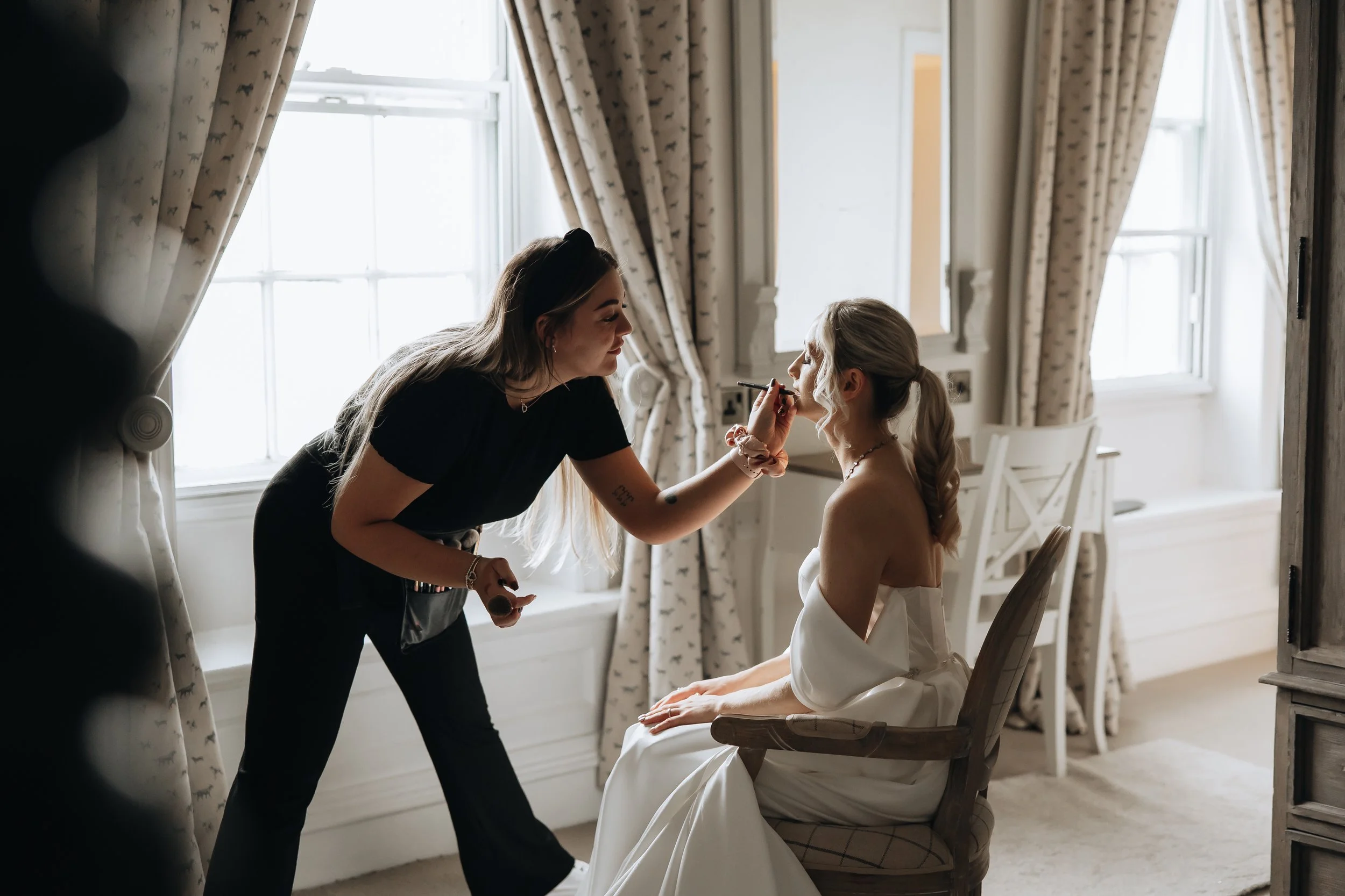 A makeup artist applying lipstick to a woman in a wedding dress, seated in a room with large windows and patterned curtains.