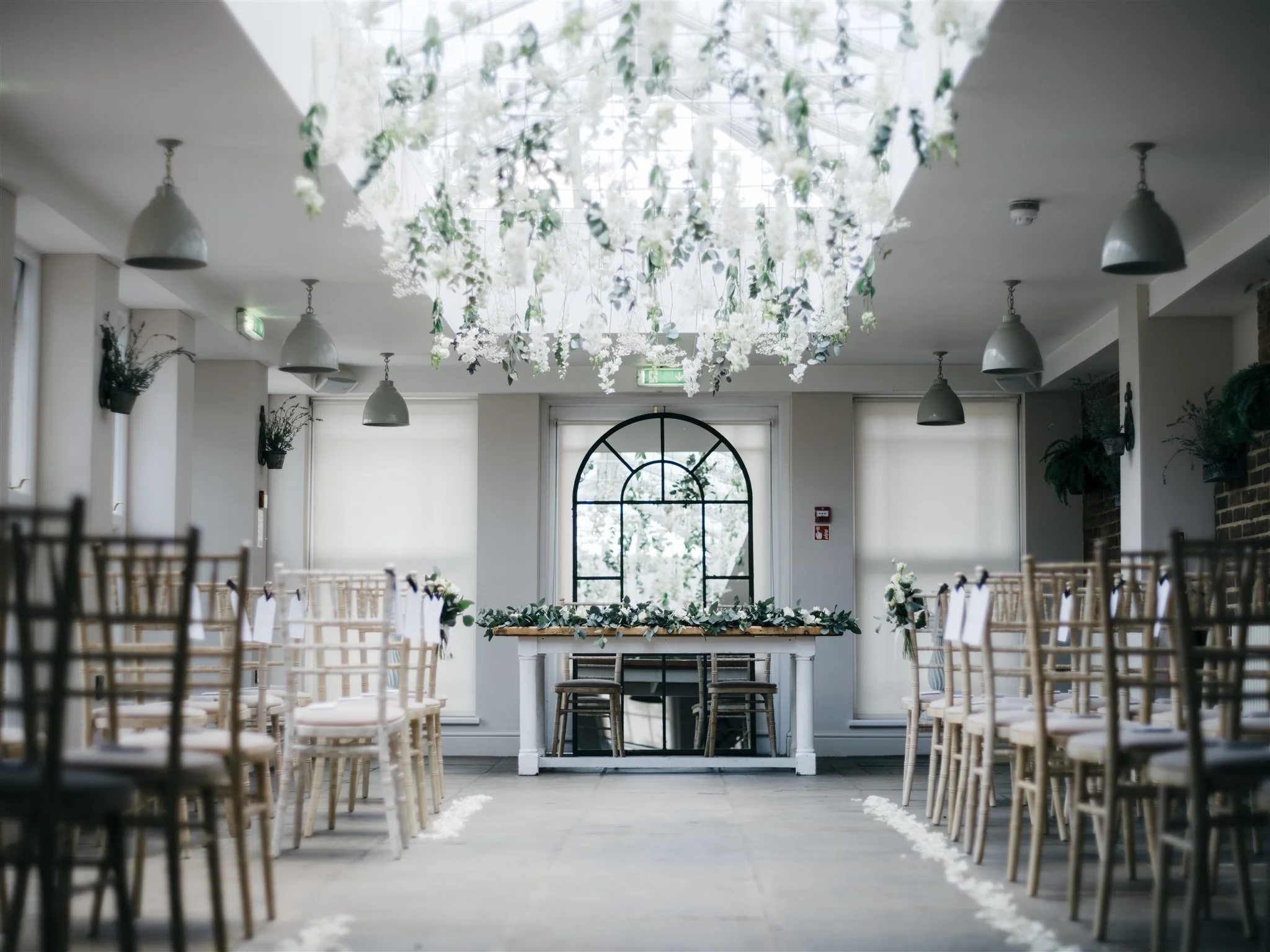 Wedding ceremony setup with rows of wooden chairs and a table decorated with greenery in a bright, modern venue with a large arched window, hanging lights, and floral decor.