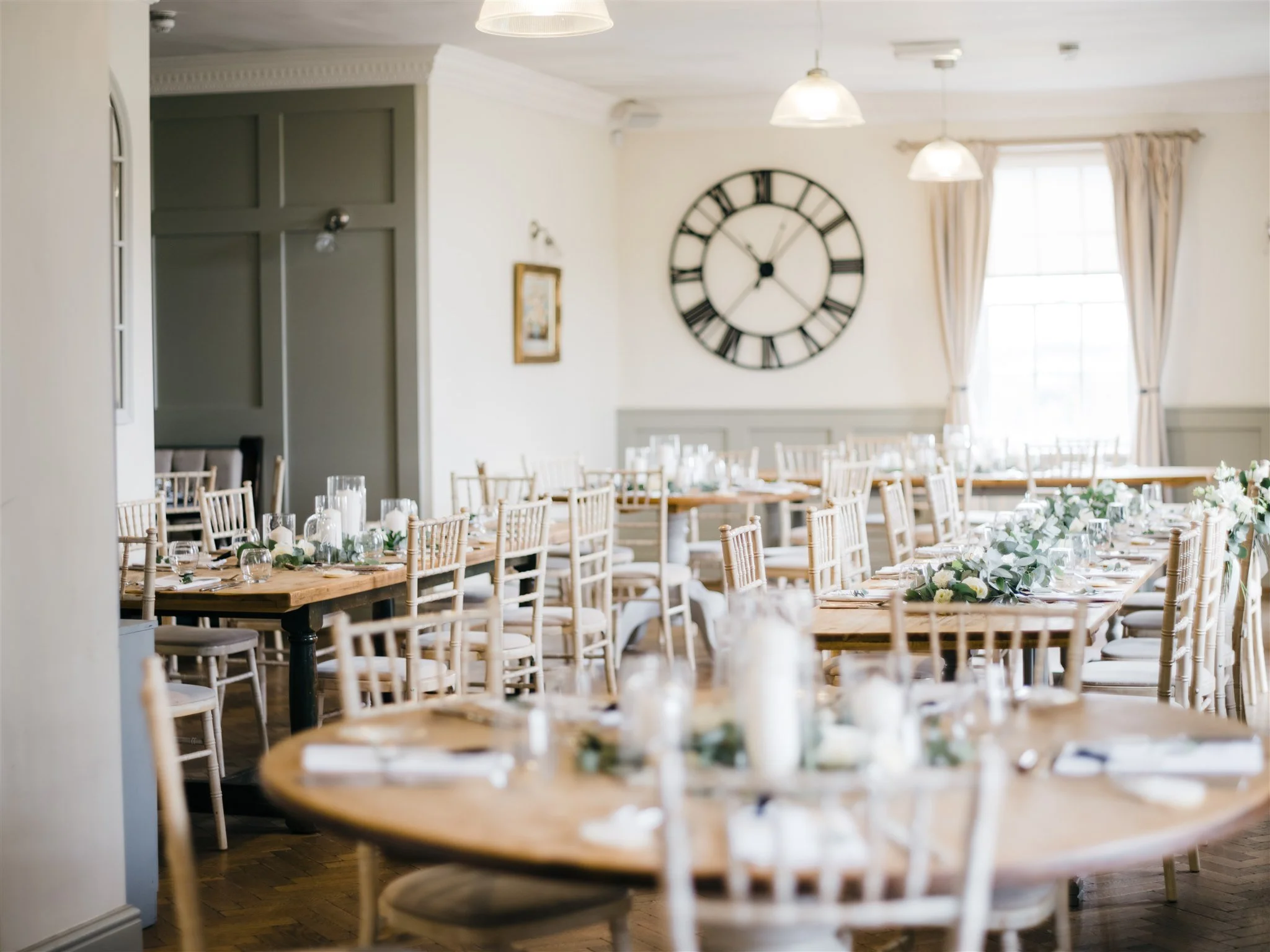 Elegantly decorated dining room with wooden tables and chairs, floral centerpieces, candles, and large windows with cream curtains, adorned with a large wall clock.